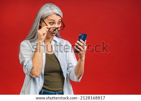 Photo portrait of asian shocked amazed surprised senior aged mature woman holding mobile phone seeing crazy discounts wearing casual isolated on red background.