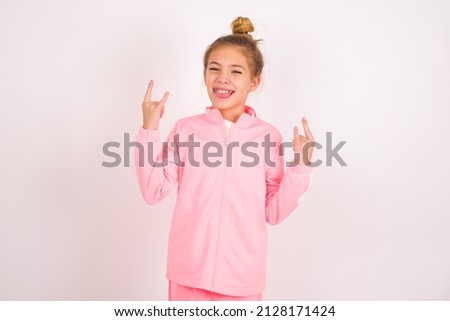 caucasian little kid girl with bun hairstyle wearing pink tracksuit over white background making rock hand gesture and showing tongue