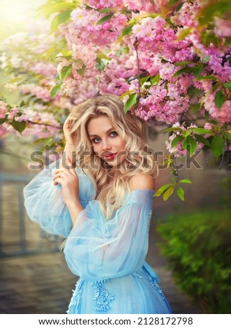 Portrait fantasy woman princess, stands near blossoming sakura tree, looks at camera, happy face pink make-up. Blonde girl goddess enjoys spring nature. Medieval vintage blue dress. Bright sunlight.