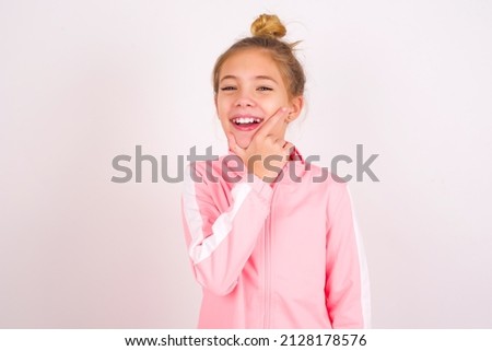 caucasian little kid girl with bun hairstyle wearing pink tracksuit over white background looking confident at the camera smiling with crossed arms and hand raised on chin. Thinking positive.