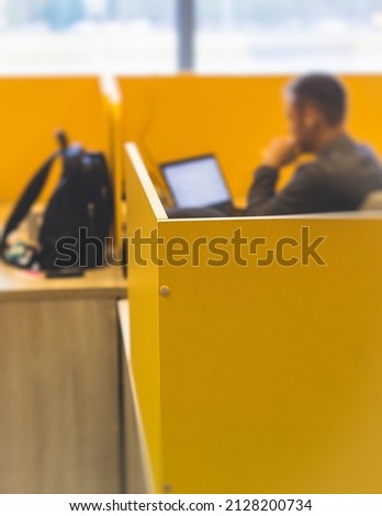 View of modern office space interior with desks in yellow tones with workers, employee with a laptop on a cozy workplace in the background, it-company open space with tables, chairs and rooms