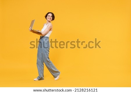 Full length side view young smiling freelancer fun woman 20s wear white tank top shirt using laptop pc computer chat online browsing internet walk isolated on yellow color background studio portrait