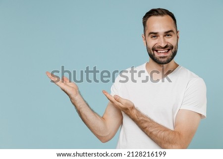 Young smiling happy fun cheerful man 20s wearing casual white t-shirt point hands arms aside on workspace area mock up copy space isolated on plain pastel light blue color background studio portrait.