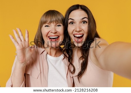Close up two young excited smiling happy daughter mother together couple women wearing casual beige clothes do selfie shot pov on mobile phone waving hands isolated on plain yellow background studio
