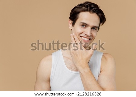 Attractive smiling calm young man he 20s perfect skin wearing bundershirt touch face after shaving isolated on light pastel beige background studio. Skin care healthcare cosmetic procedures concept. Royalty-Free Stock Photo #2128216328