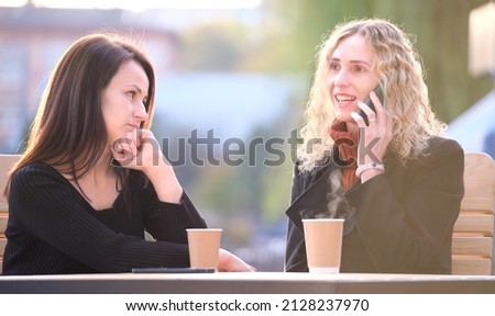 Sad woman being ignored by her friend sitting at street cafe outdoors while she is talking happily on mobile phone and paying no attention. Friendship problems concept Royalty-Free Stock Photo #2128237970
