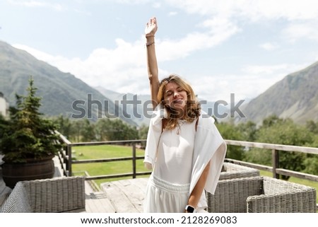 Satisfied excited smiling girl with wavy light hair wearing white outfit is holding up hand with closed eyes and enjoying travelling in mountains. High quality photo