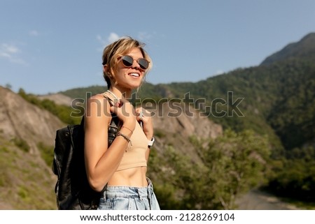 Happy smiling girl with wavy hair wearing t-shirt with backpack traveling in mountains in sunlight. Curly lady in and stylish summer outfit moves on mountains background. High quality photo