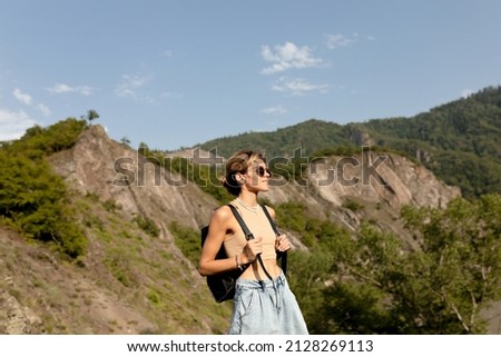 Adorable charming lady with wavy light hair wearing t-shirt and shorts with backpack is moving against green mountains in sunlight in warm spring day. Woman traveling in Woman's Day. 