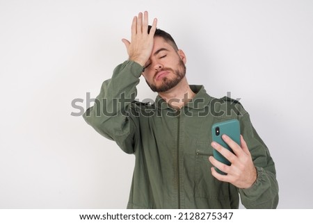 Upset depressed young caucasian man wearing working overalls over white background makes face palm as forgot about something important holds mobile phone expresses sorrow and regret blames