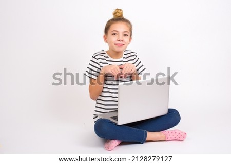 Beautiful caucasian teen girl sitting with laptop in lotus position on white background makes bunny paws and looks with innocent expression plays with her little kid