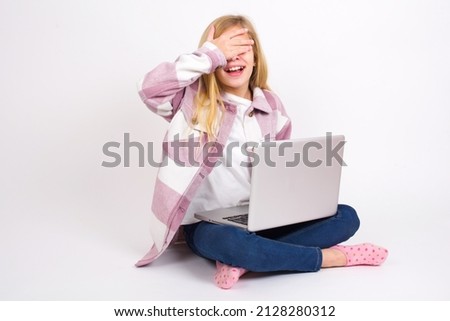 Happy Beautiful caucasian teen girl sitting with laptop in lotus position on white background closing eyes with hand going to see surprise prepared by friend standing and smiling in anticipation 