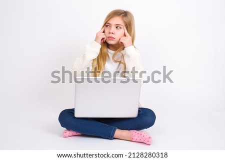 Beautiful caucasian teen girl sitting with laptop in lotus position on white background with thoughtful expression, looks away, keeps hand near face, thinks about something pleasant.