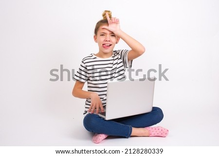 Beautiful caucasian teen girl sitting with laptop in lotus position on white background gestures with finger on forehead makes loser gesture makes fun of people shows tongue