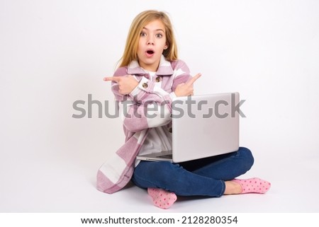 Confused Beautiful caucasian teen girl sitting with laptop in lotus position on white background chooses between two ways, points at both sides with crossed hands, feels doubt. Need your advice.