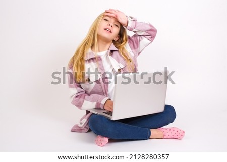 Beautiful caucasian teen girl sitting with laptop in lotus position on white background wiping forehead with hand making phew gesture, expressing relief feels happy that he prevented huge disaster. 
