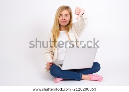 Discontent Beautiful caucasian teen girl sitting with laptop in lotus position on white background shows disapproval sign, keeps thumb down, expresses dislike, frowns face in discontent. Negative 