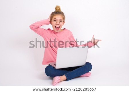 Surprised Beautiful caucasian teen girl sitting with laptop in lotus position on white background pointing at empty space holding hand on head