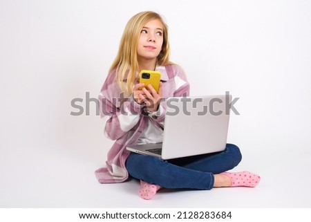Happy Beautiful caucasian teen girl sitting with laptop in lotus position on white background listening to music with earphones using mobile phone.