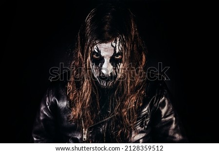 Portrait photo of black metal metalhead man with long brown hair and painted face standing in leather jacket and looking at camera on black background.