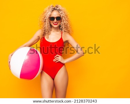 Young beautiful smiling woman posing near yellow wall in studio.Sexy model in red swimwear bathing suit.Positive female with curls hairstyle. Holding penny inflatable ball.Happy and cheerful Royalty-Free Stock Photo #2128370243