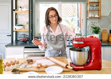 Woman wearing apron baking cookies in a cozy kitchen. The housewife makes the dough for the cake in the planetary mixer. Homemade cakes or pies. Royalty-Free Stock Photo #2128381307