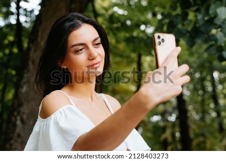 Pretty young mixed race woman taking photo, selfie portrait on mobile phone camera outdoors. Smiling brunette indian lady in white summer dress using smartphone.