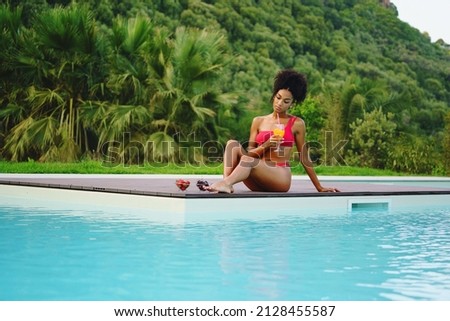 African American woman relaxing poolside drinking healthy smoothie and eating some fruit against a green hill in the summer