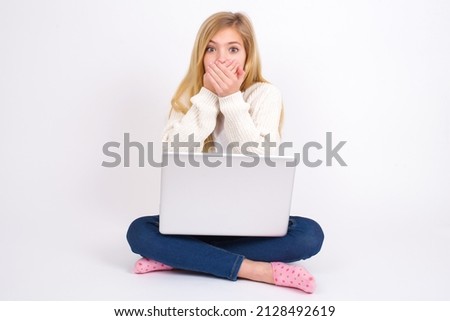 Upset Beautiful caucasian teen girl sitting with laptop in lotus position on white background, covering her mouth with both palms to prevent screaming sound, after seeing or hearing something bad.