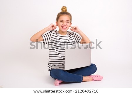 Happy Beautiful caucasian teen girl sitting with laptop in lotus position on white background ignores loud music and plugs ears with fingers asks to turn off sound