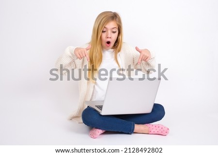 Amazed Beautiful caucasian teen girl sitting with laptop in lotus position on white background points down with fore fingers, opens mouth being shocked. Advertisement concept.