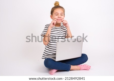 Pleased Beautiful caucasian teen girl sitting with laptop in lotus position on white background with closed eyes keeps hands near cheeks and smiles tenderly imagines something very pleasant