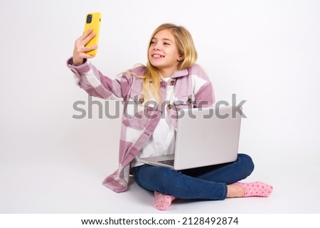 Isolated shot of pleased cheerful Beautiful caucasian teen girl sitting with laptop in lotus position on white background , makes selfie with mobile phone. People, technology and leisure concept