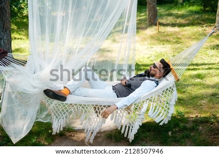 A bearded man in sunglasses and a suit  lies in a hammock outdoors.
