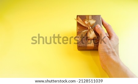 A man's hand holding brown gift box with gold ribbon isolated on a yellow background. Valentine concept, christmas and newyear concept. Anniversary and Thanksgiving concept.