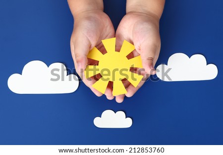 Child holds in his palms a paper sun in the blue sky with paper clouds.
