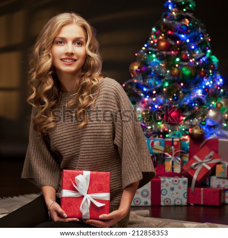 young happy smiling casual woman holding red gift over christmas tree and lights on background. warm light