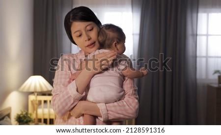 waist up asian mom is embracing and patting on her baby’s back trying to put her to sleep at nighttime in illuminated home interior. Royalty-Free Stock Photo #2128591316