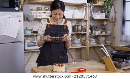 cheerful asian young girl running a small home baking business by kitchen table and using phone to take pictures for her product in a wooden style kitchen