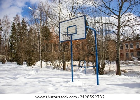 An old basketball white and blue shield with a basketball ring outdoors in the countryside on a sunny day. Lots of snow, trees and a blue sky with fluffy clouds. Healthy lifestyle.