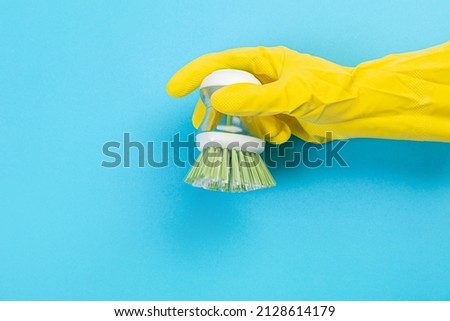 A hand in a yellow glove holds a plastic cleaning brush on a blue background. Banner with copy space. Chemical cleaners, household chemicals, brushes and collage supplies. 