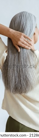 Middle aged woman in yellow blouse adjusts long loose hoary hair on light grey background in studio backside view. Mature beauty lifestyle