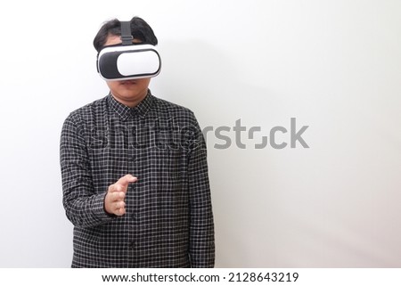 Portrait of Asian man in black plaid shirt using Virtual Reality (VR) glasses reaching out for a handshake and greet someone or his friend inside the game. Isolated image on white background