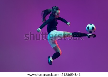 In action. One sportive girl, female soccer player training with football ball isolated on purple studio background in neon light. Concept of sport, action, motion, fitness. Young sportive girl in Royalty-Free Stock Photo #2128654841