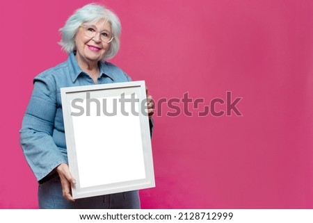grey-haired senior woman holding empty picture frame on pink background