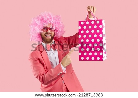 Funny crazy clown man in pink curly wig, sunglasses, funky suit and bow tie showing pink and white polka dot paper bag and smiling. Shopping, sale, presents, discounts, special offers concept Royalty-Free Stock Photo #2128713983