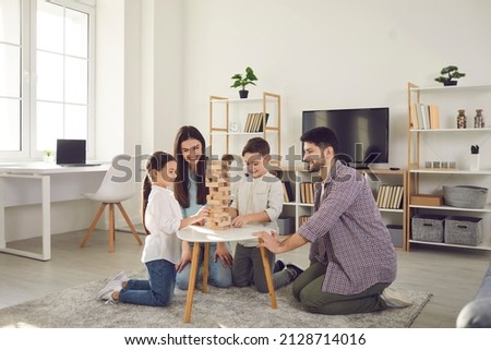 Happy family with kids playing board games at home. Smiling mom, dad and children sitting on the floor around a low table in a cozy living room and pulling wooden blocks from the tower Royalty-Free Stock Photo #2128714016