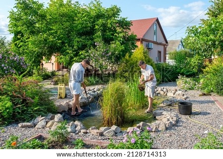 Mature father and young adult son cleaning artificial garden pond bottom with high-pressure washer nozzle and landing net from mud and sludge. Spring and summer pond care work