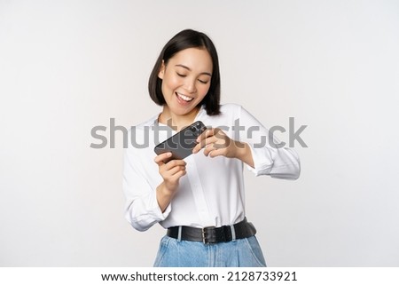 Young korean woman, asian girl playing mobile video game on smartphone, looking at horizontal phone screen, standing over white background