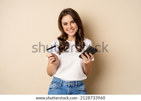 Image of young happy woman paying online, holding smartphone and credit card, enter info in application on mobile phone, standing against beige background